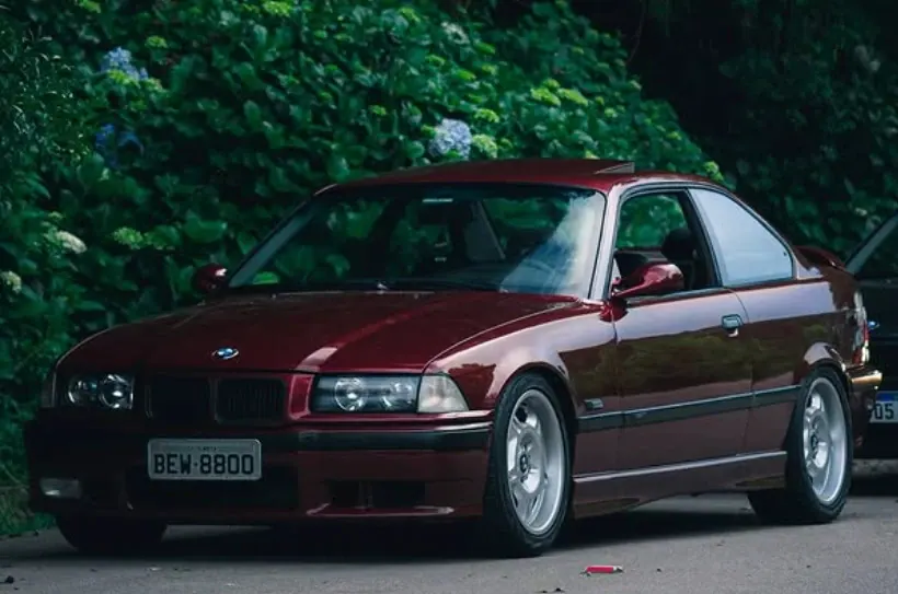 BMW E36 coupé vinho com rodas prata, estacionada diante de hortênsias